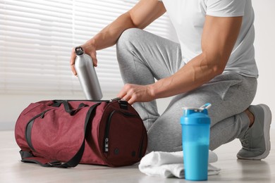 Young man putting bottle of water into bag indoors, closeup. Shaker with protein and towel on floor