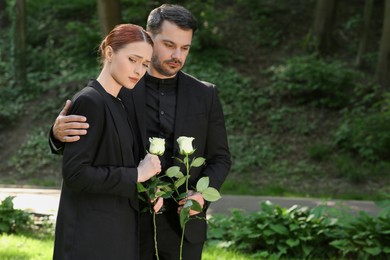 Sad couple with white rose flowers mourning outdoors, space for text. Funeral ceremony