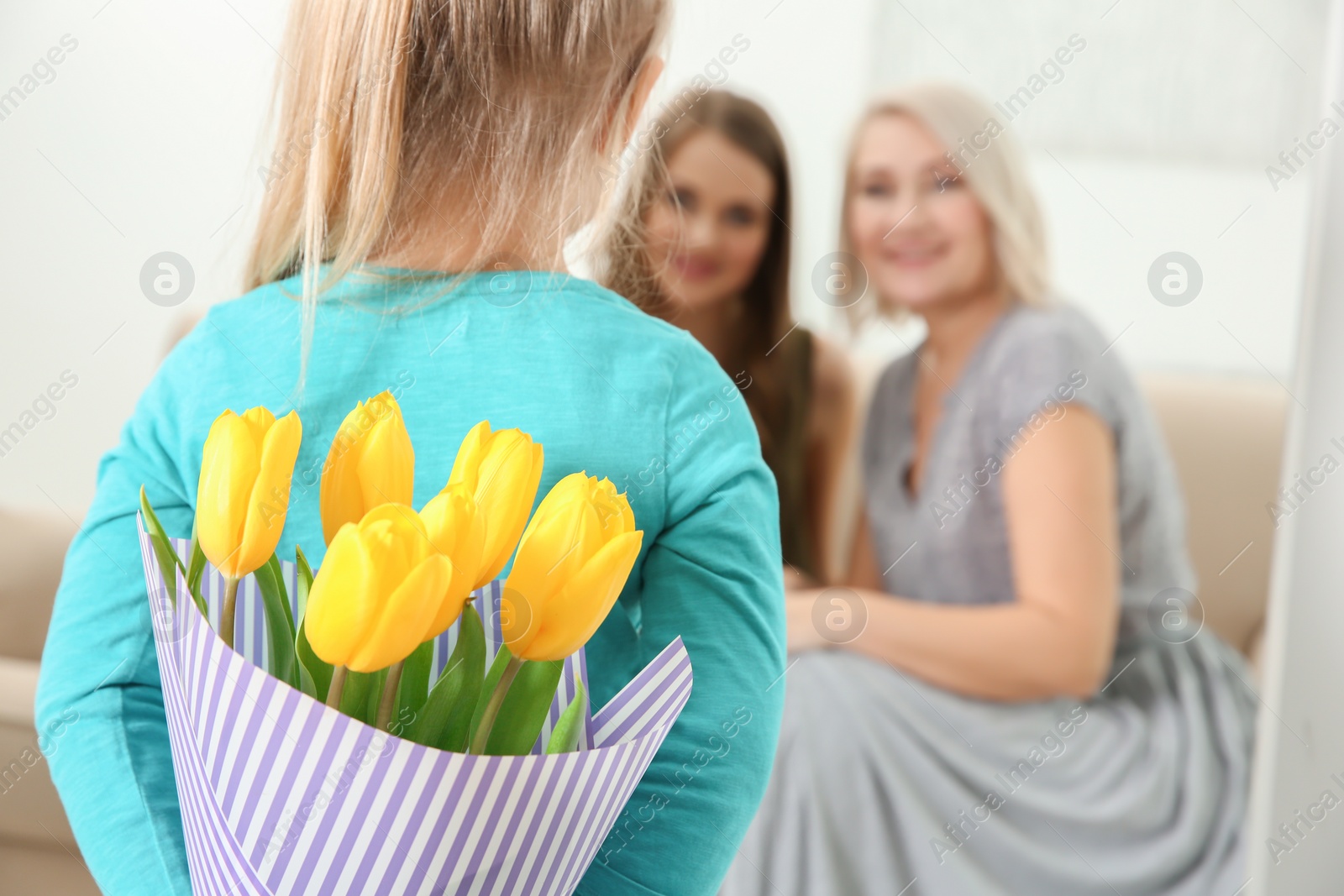 Photo of Little girl hiding tulip bouquet for mother and grandmother behind her back at home