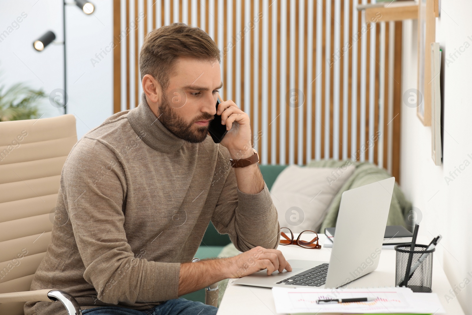 Photo of Young man talking on phone while working with laptop at home