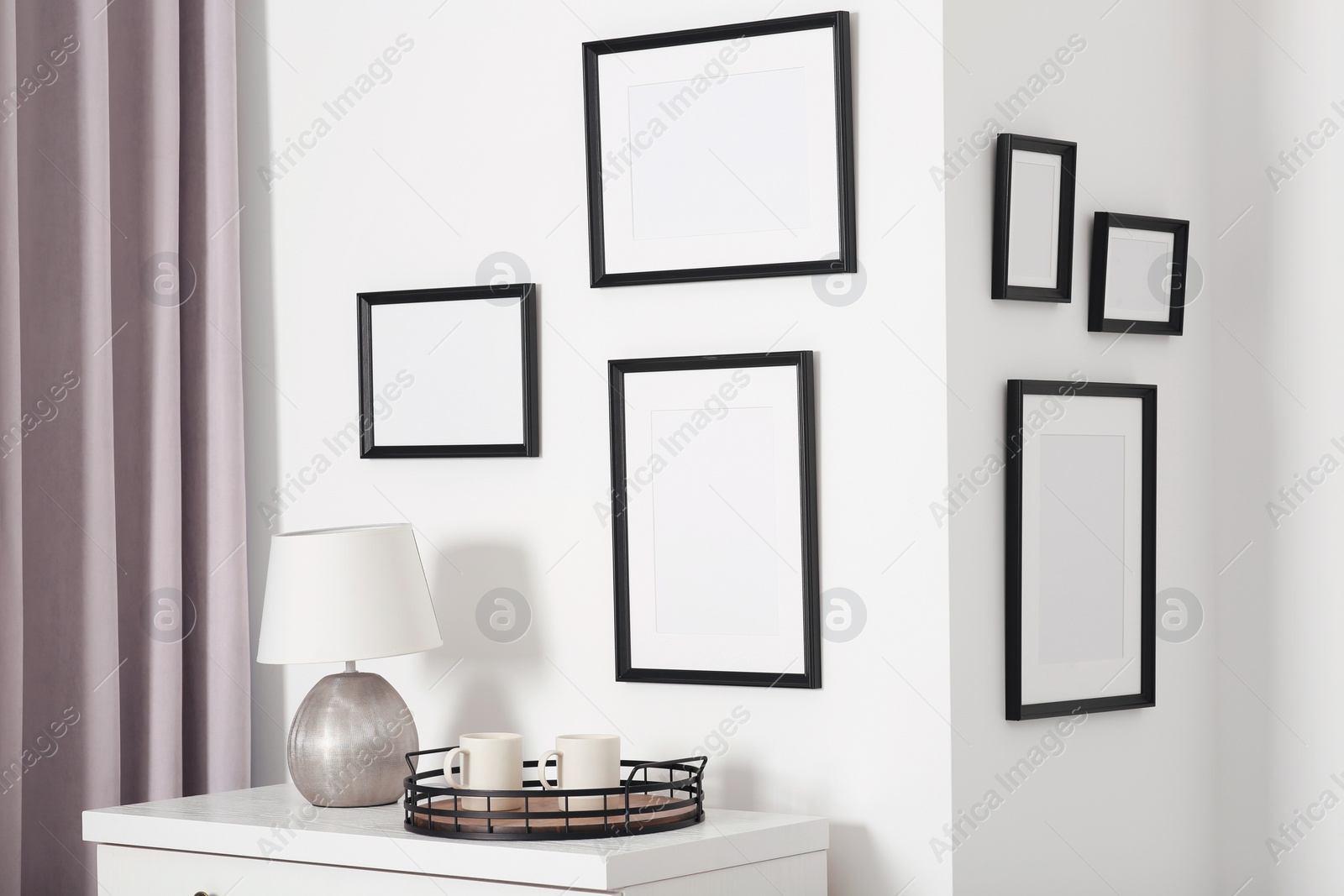 Photo of Empty frames hanging on white wall, cups and lamp indoors