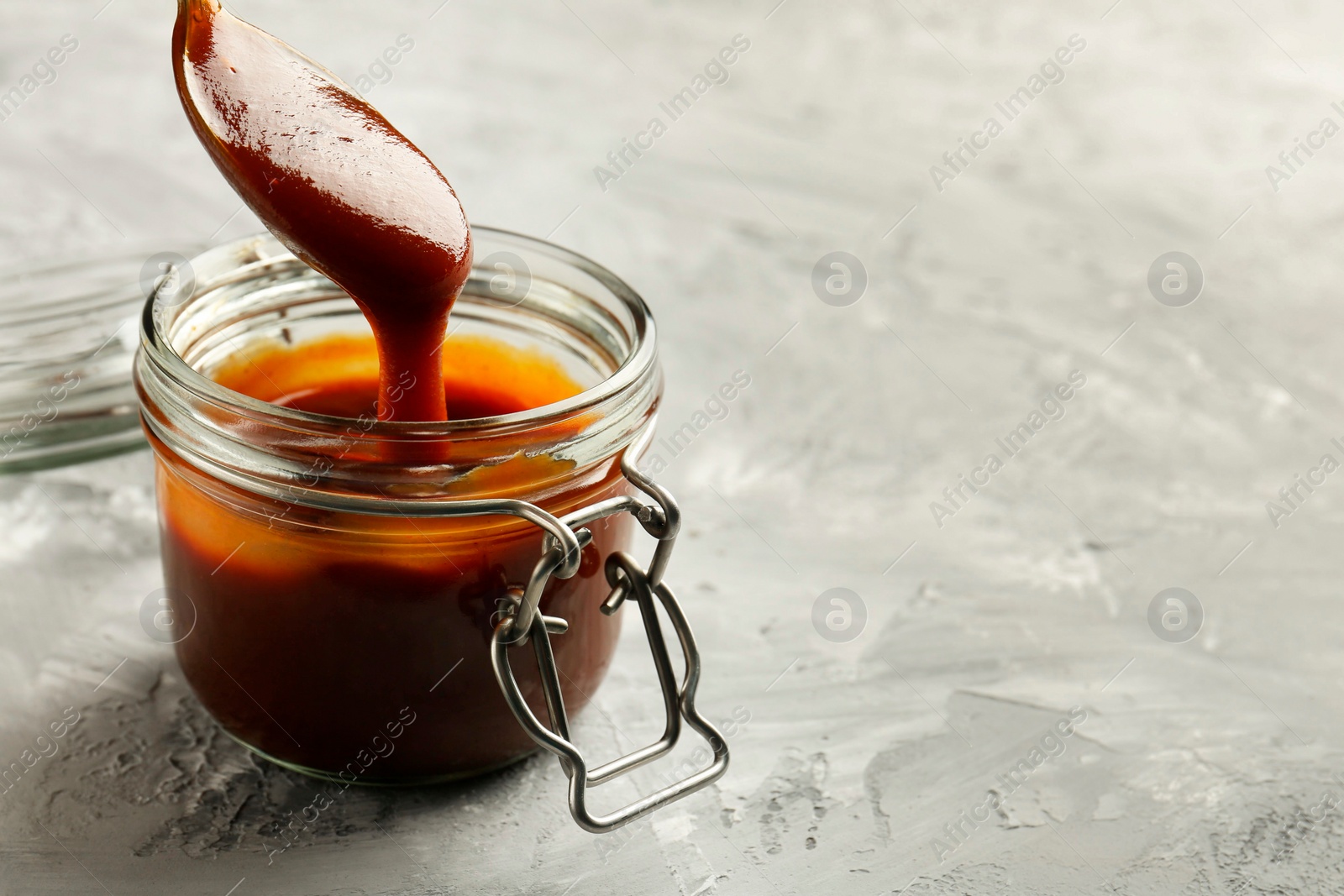 Photo of Taking tasty barbeque sauce from jar with spoon at grey textured table, closeup. Space for text