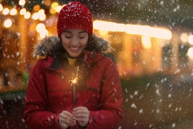 Image of Happy young woman with sparkler at winter fair in evening. Christmas celebration