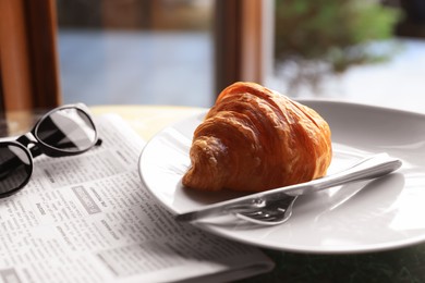 Photo of Tasty croissant, newspaper and sunglasses on table in cafeteria