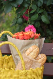 Beautiful bouquet of roses and baguettes in yellow wicker bag on bench outdoors