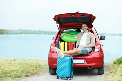 Happy man sitting in car trunk with suitcases on riverside. Space for text