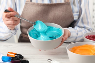 Woman mixing cream with light blue food coloring at white wooden table, closeup