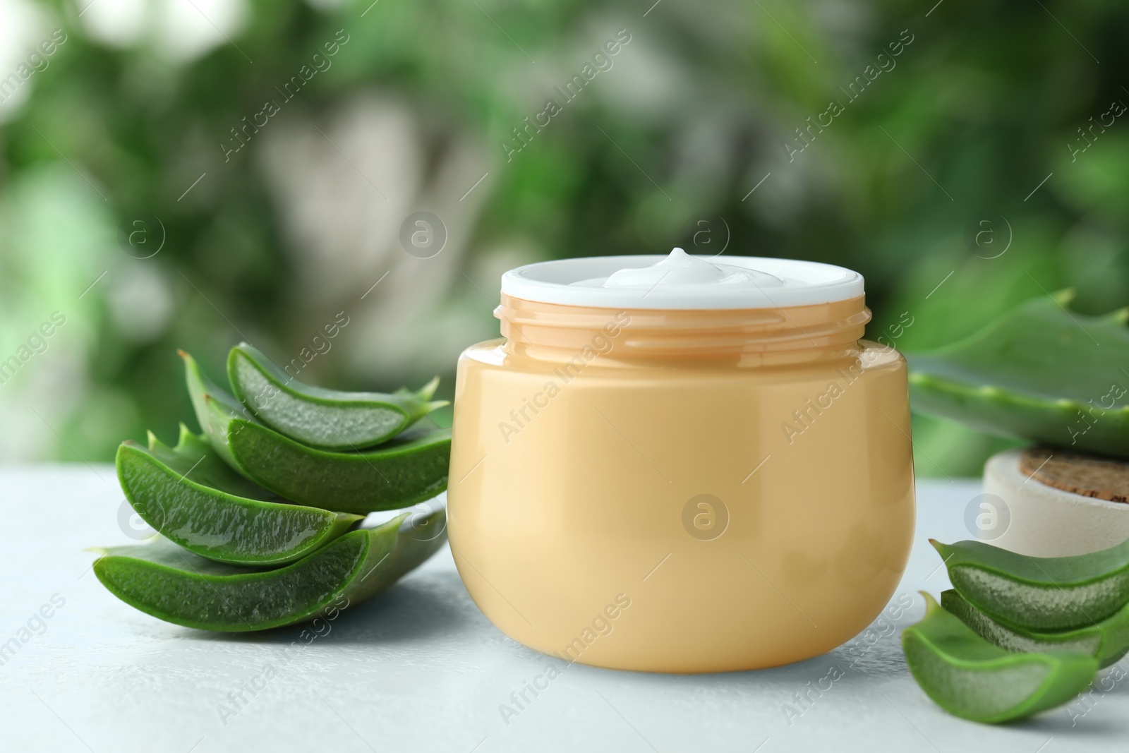 Photo of Jar with cream and cut aloe leaf on white table against blurred green background, closeup