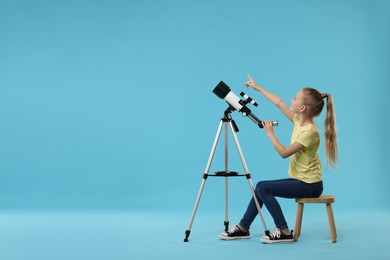 Photo of Excited little girl with telescope pointing at something on light blue background, space for text
