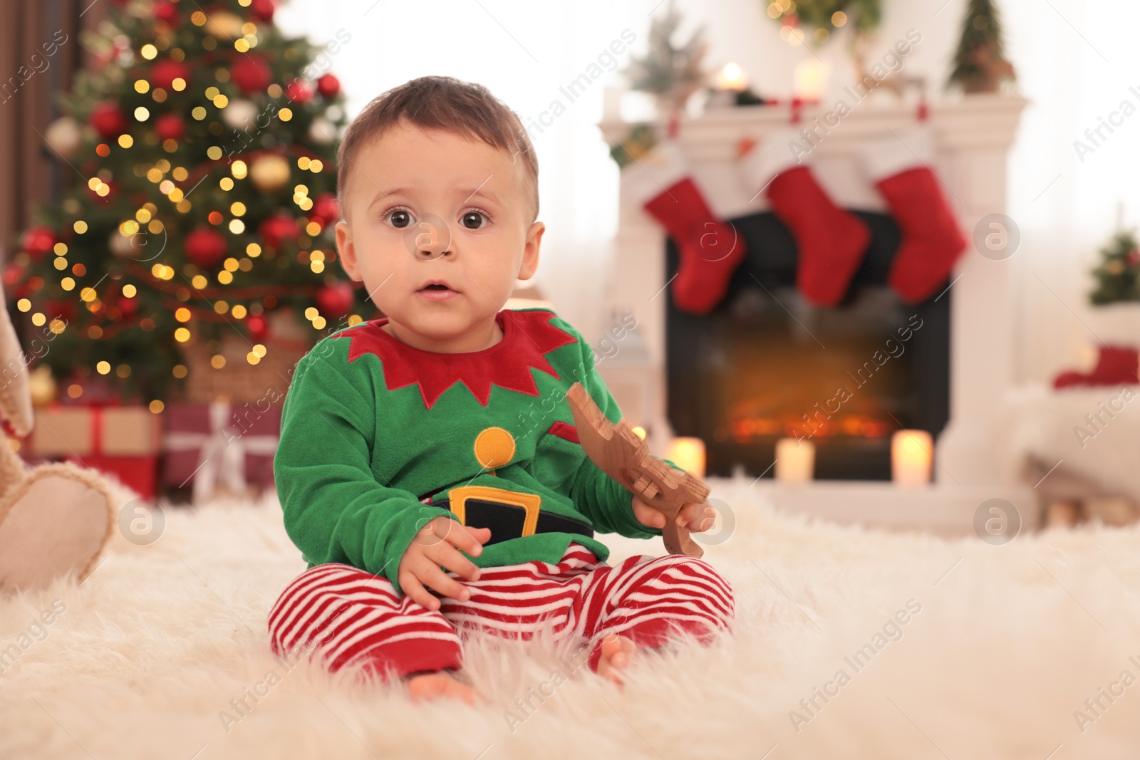 Photo of Baby wearing cute elf costume on floor in room decorated for Christmas