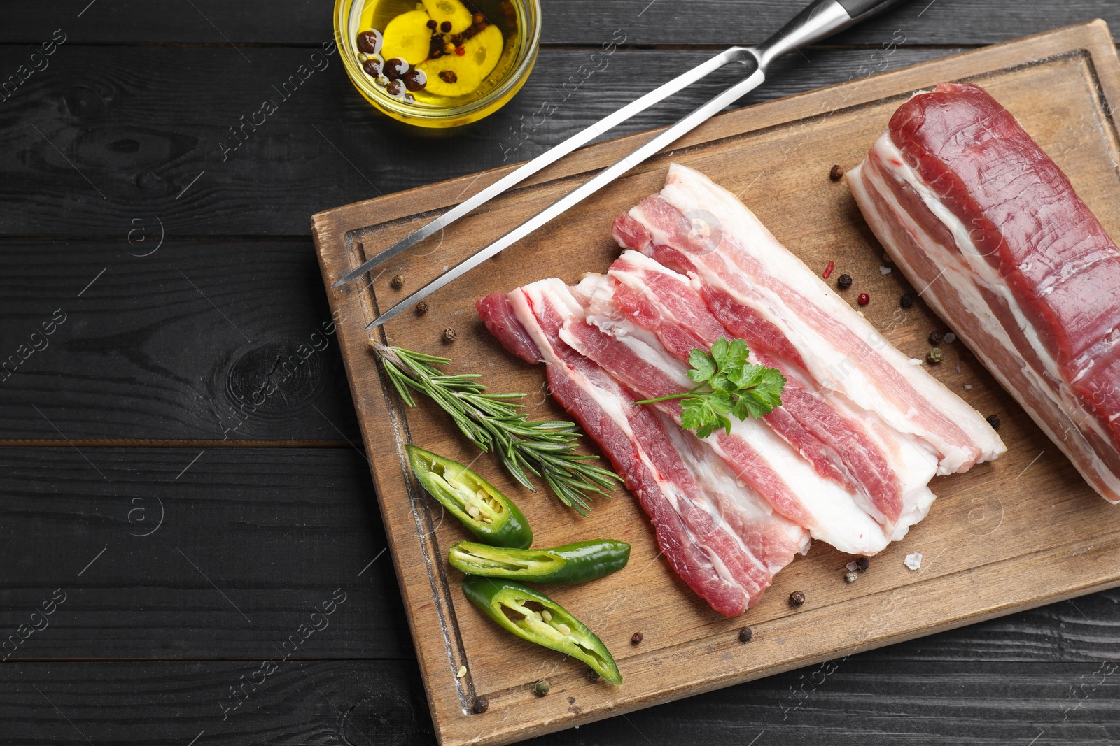 Photo of Pieces of raw pork belly, chili pepper, peppercorns, rosemary and parsley on black wooden table, flat lay