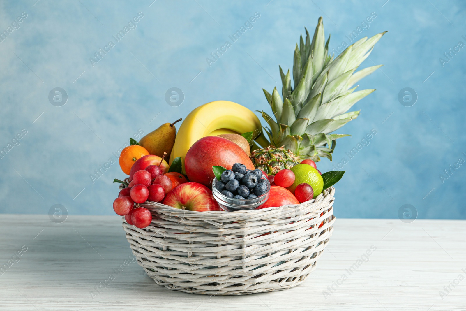 Photo of Assortment of fresh exotic fruits in wicker bowl on white wooden table against light blue background