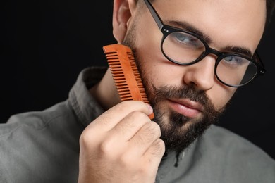 Photo of Handsome young man combing beard on black background, closeup