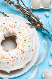 Photo of Delicious Easter cake decorated with sprinkles near painted eggs and willow branches on light blue background, flat lay