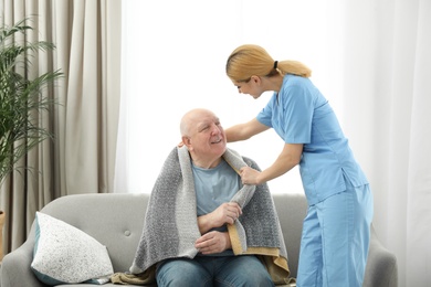 Nurse covering elderly man with blanket indoors. Assisting senior people