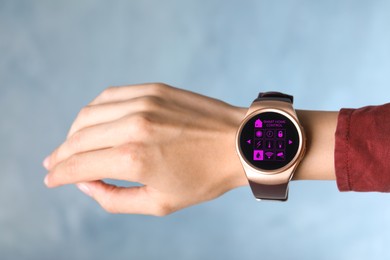 Woman wearing smartwatch with icons of smart home control app on display against light blue background, closeup