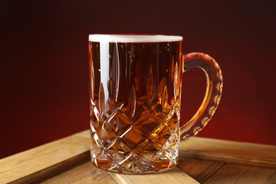 Photo of Mug with fresh beer on wooden crate against burgundy background, closeup