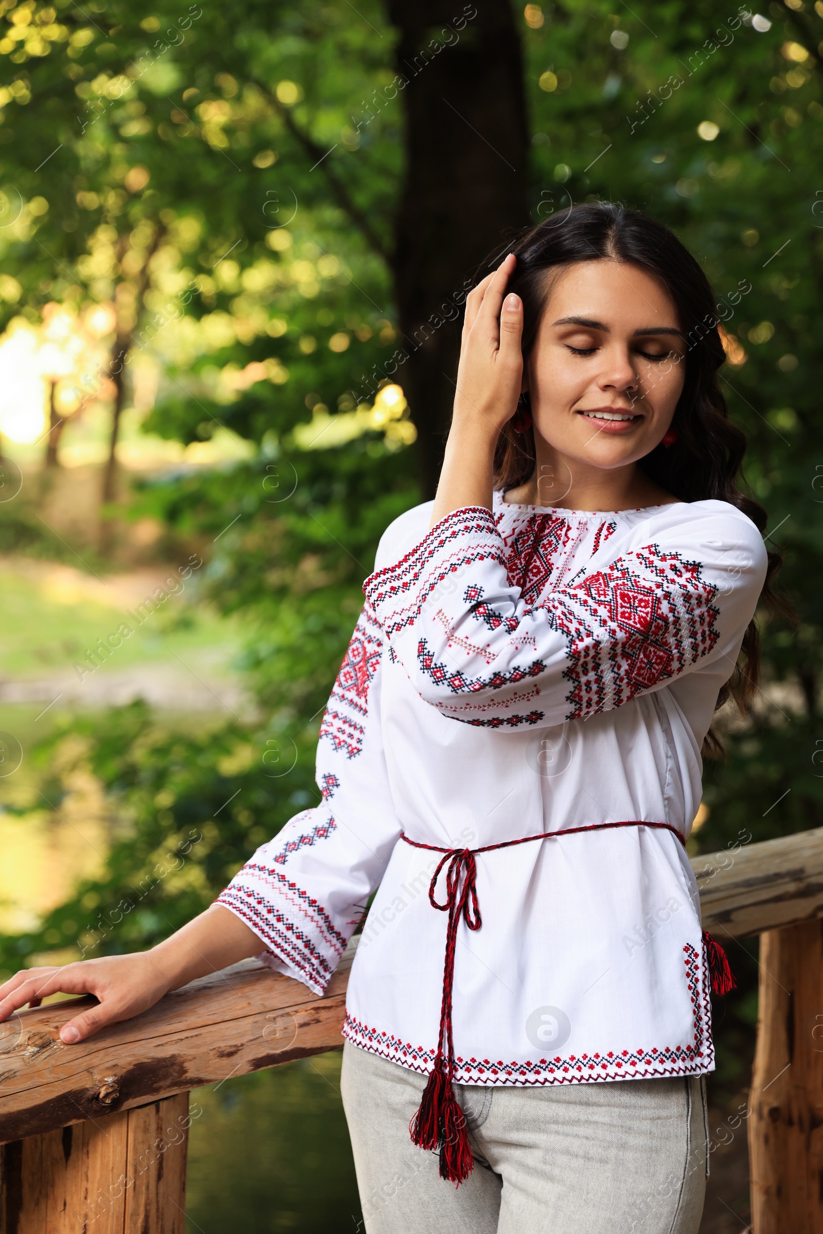 Photo of Beautiful woman wearing embroidered shirt near wooden railing in countryside. Ukrainian national clothes