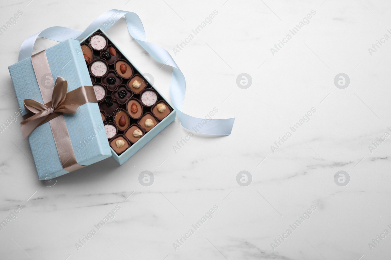 Photo of Open box of delicious chocolate candies and light blue ribbon on white marble table, flat lay. Space for text