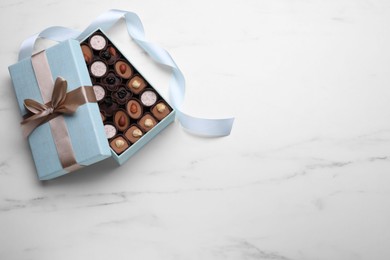 Open box of delicious chocolate candies and light blue ribbon on white marble table, flat lay. Space for text