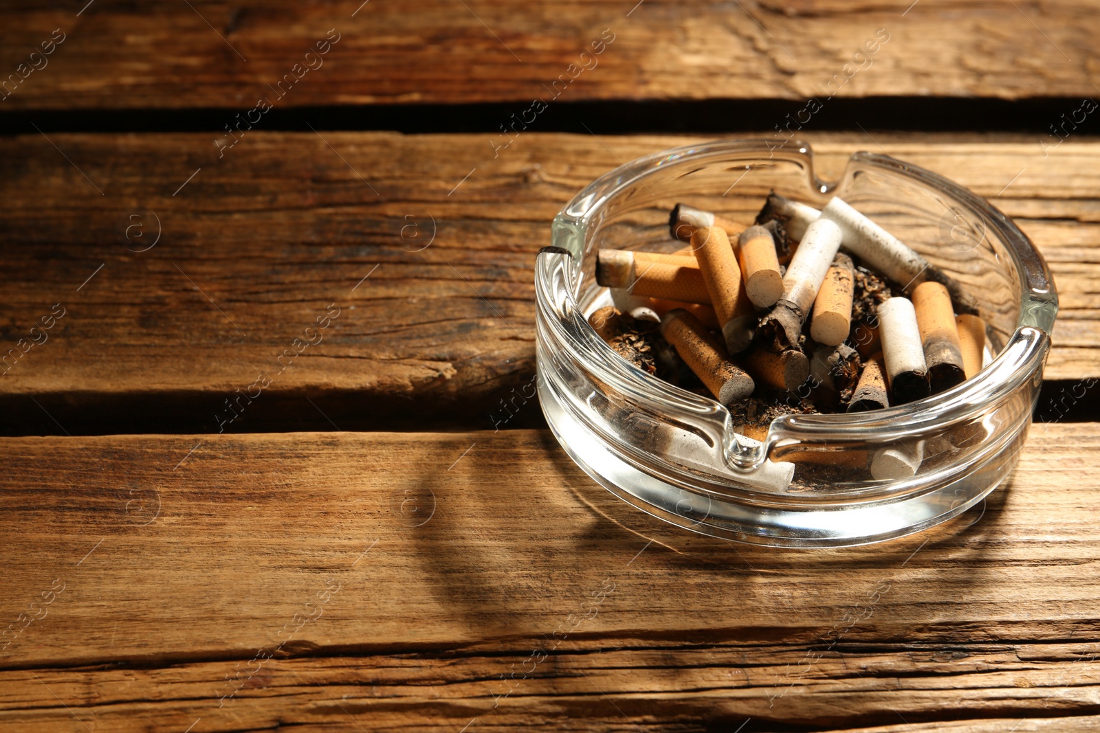 Photo of Glass ashtray with cigarette stubs on wooden table. Space for text