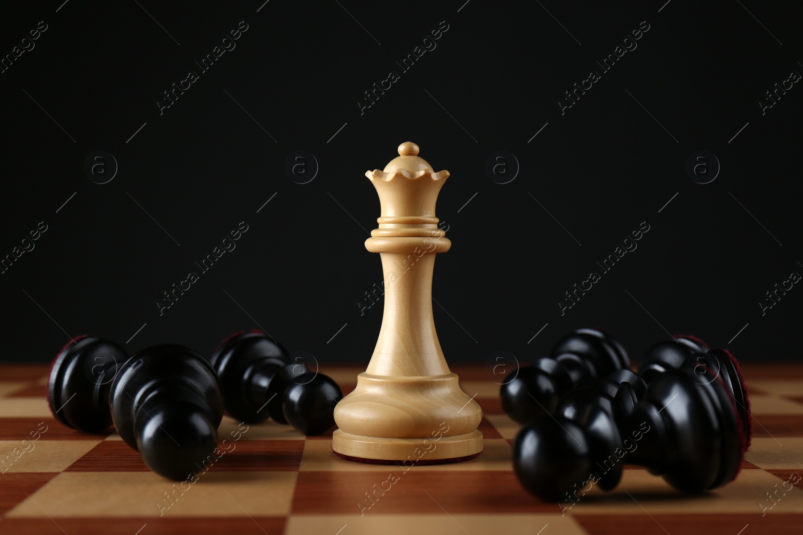 Photo of White queen among fallen black pawns on wooden chess board against dark background