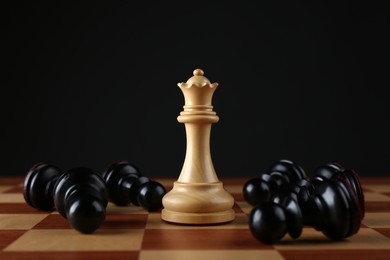 Photo of White queen among fallen black pawns on wooden chess board against dark background