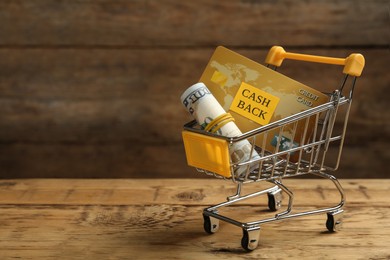 Photo of Shopping cart with rolled dollar banknotes and credit card on wooden table, space for text. Cashback concept