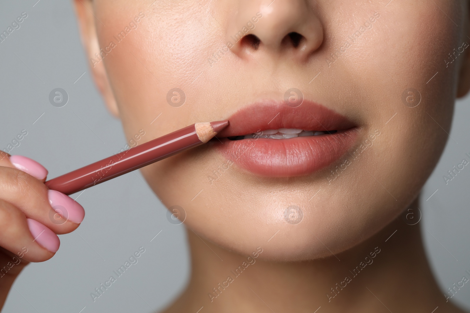 Photo of Young woman applying beautiful nude lip pencil on grey background, closeup