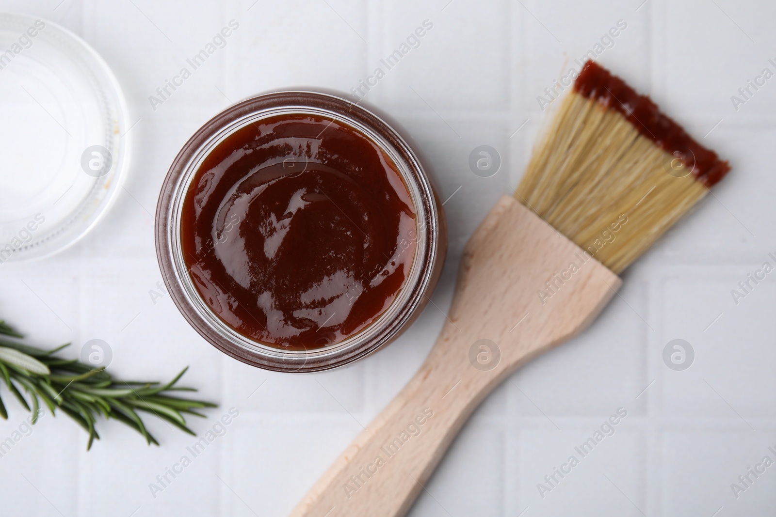 Photo of Marinade in jar, rosemary and basting brush on white tiled table, flat lay