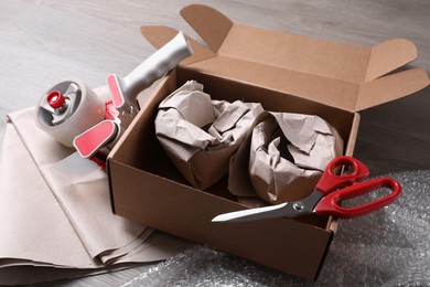 Photo of Open box with wrapped items, adhesive tape, scissors, paper and bubble wrap on wooden table