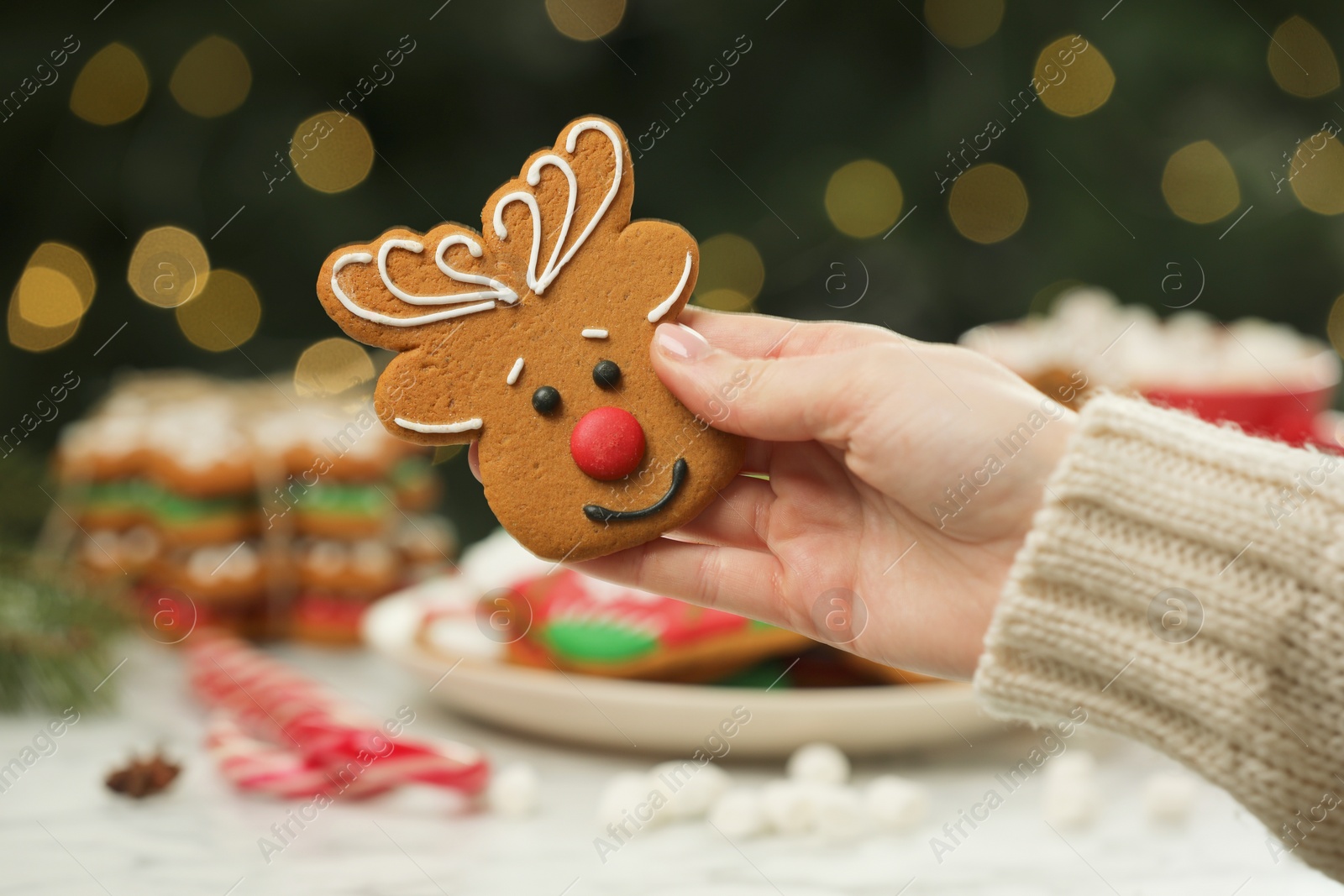 Photo of Woman with decorated cookie at table against blurred Christmas lights, closeup