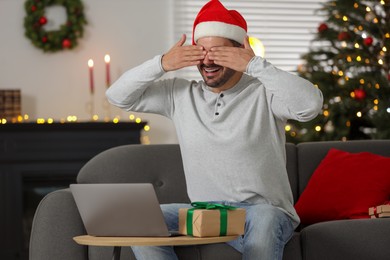 Celebrating Christmas online with exchanged by mail presents. Man covering eyes before opening gift box during video call on laptop at home