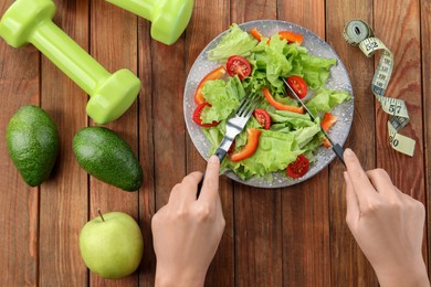 Photo of Healthy diet. Woman eating salad at wooden table with measuring tape and dumbbells, top view