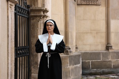 Young nun with hands clasped together praying near metal door outdoors