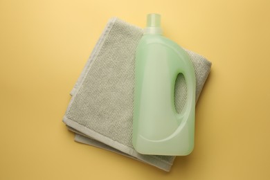 Photo of Bottle of fabric softener and towel on pale yellow background, top view