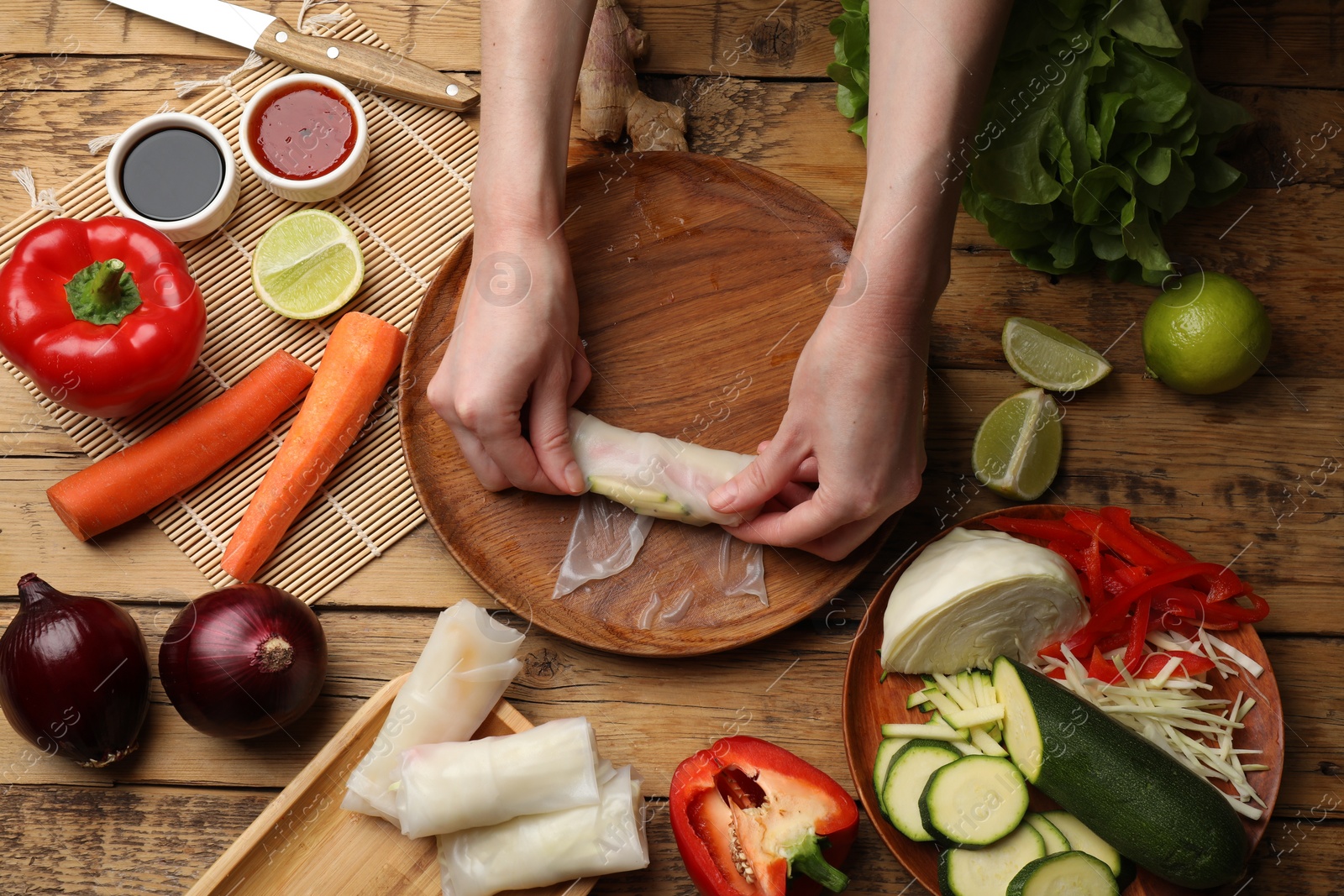 Photo of Making delicious spring rolls. Woman wrapping fresh vegetables into rice paper at wooden table, flat lay