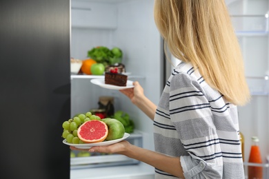 Photo of Choice concept. Woman taking plates with fruits and cake from refrigerator in kitchen, closeup