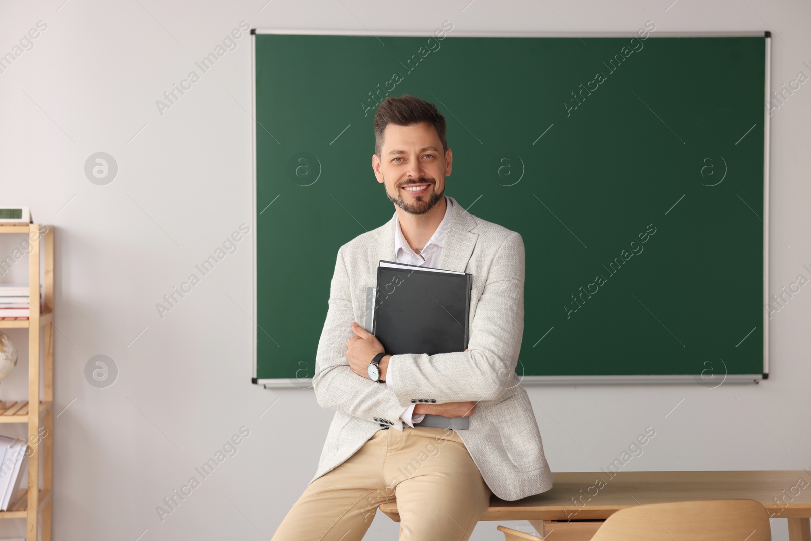 Photo of Happy teacher with stationery at desk in classroom