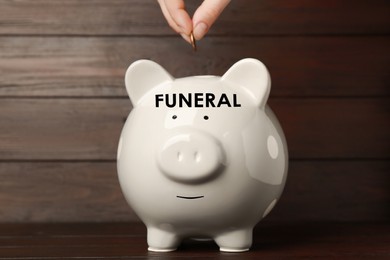 Money for funeral expenses. Woman putting coin into white piggy bank at wooden table, closeup