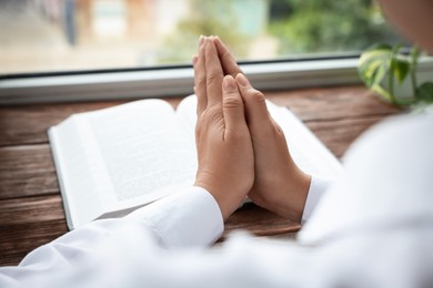 Woman holding hands clasped while praying at wooden table with Bible, closeup