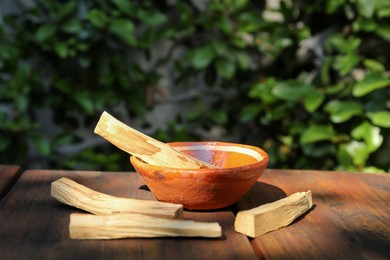 Photo of Palo santo sticks and bowl on wooden table outdoors, space for text