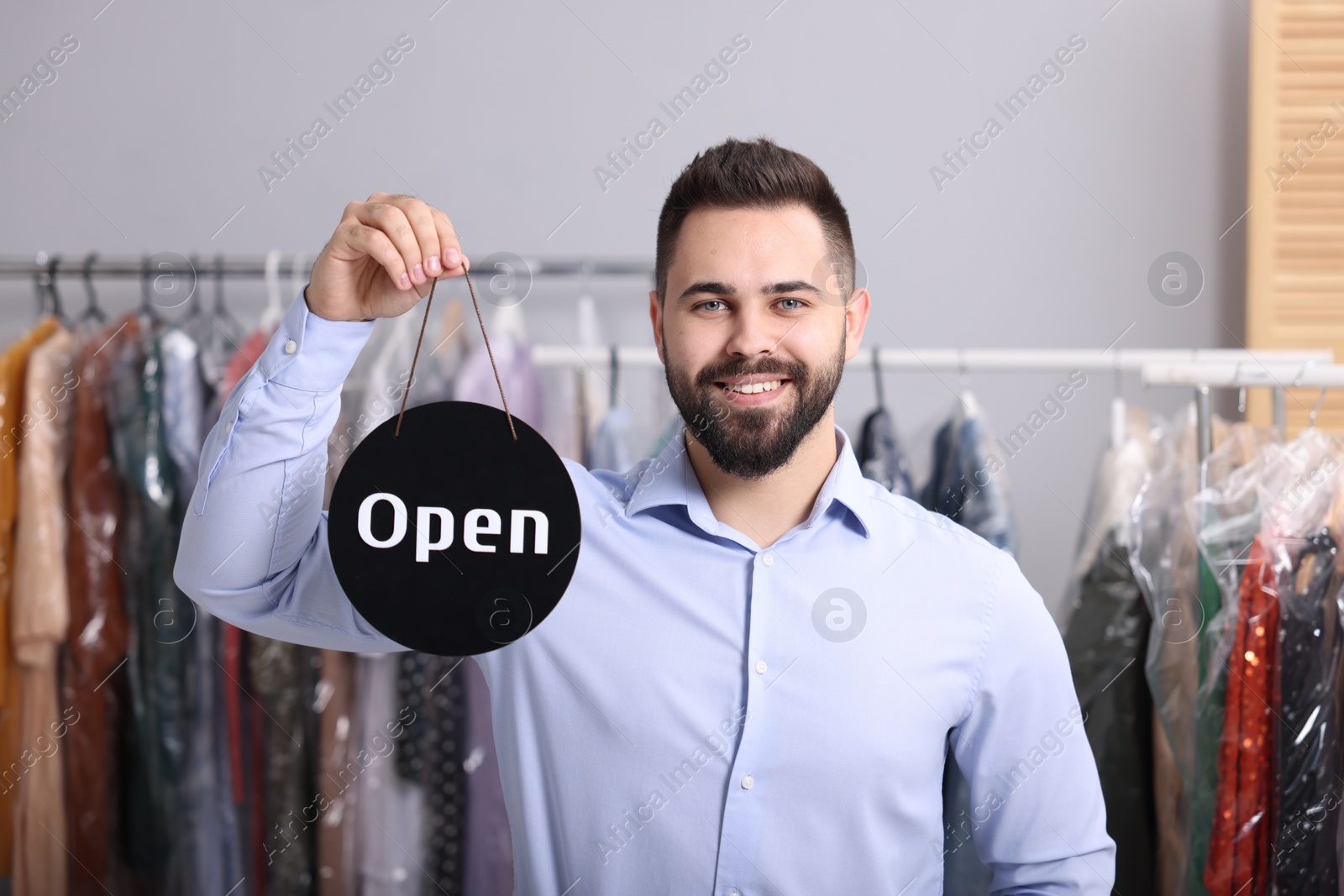 Photo of Dry-cleaning service. Happy worker holding Open sign indoors