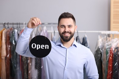 Photo of Dry-cleaning service. Happy worker holding Open sign indoors