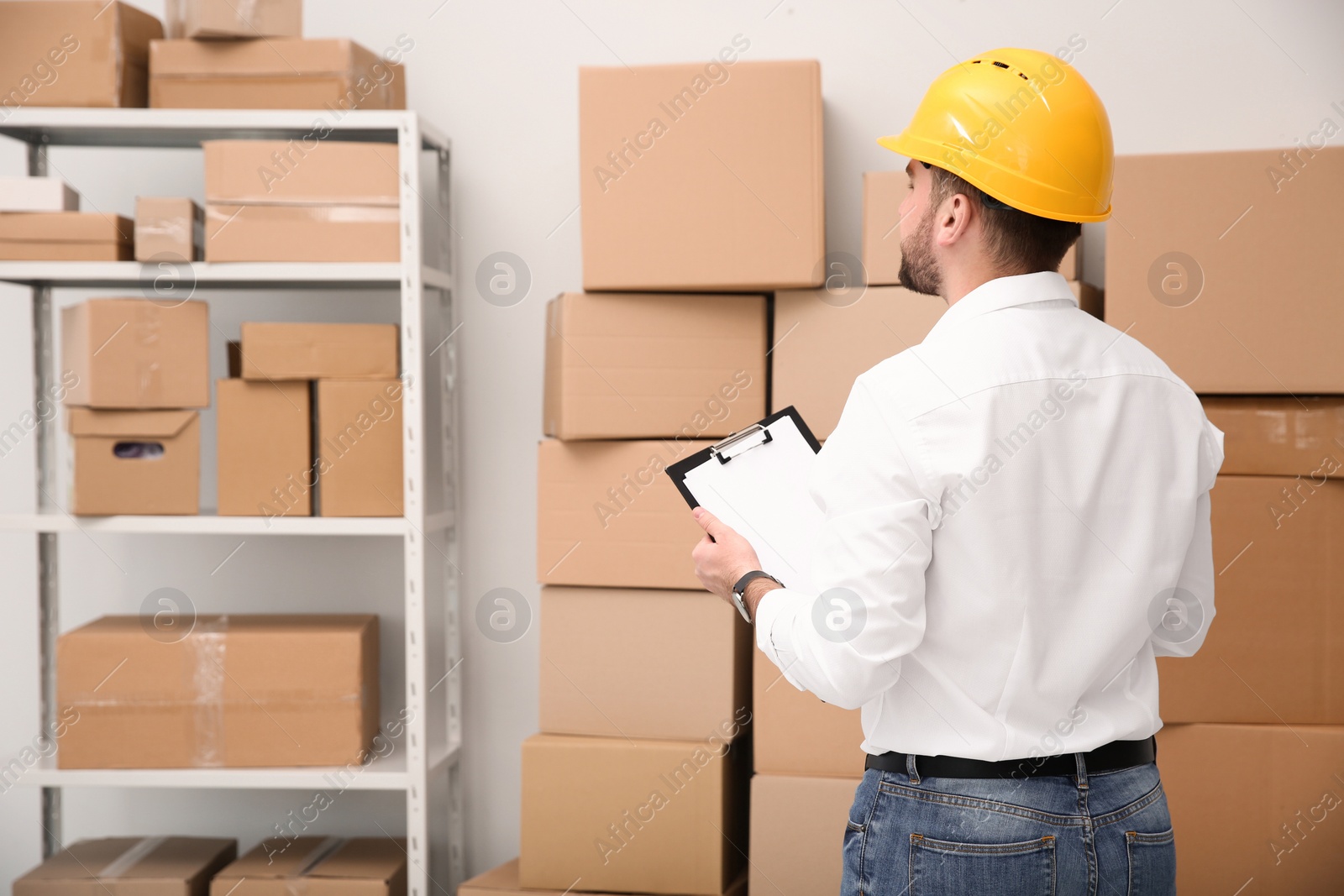 Photo of Young man with clipboard near cardboard boxes at warehouse, back view