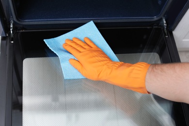 Photo of Young man cleaning oven with rag in kitchen, closeup