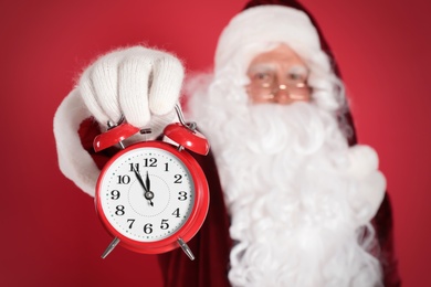 Photo of Santa Claus holding alarm clock on red background, focus on hand. Christmas countdown