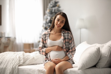 Happy pregnant woman on bed in room decorated for Christmas. Expecting baby