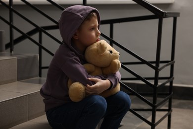 Child abuse. Upset boy with teddy bear sitting on stairs indoors
