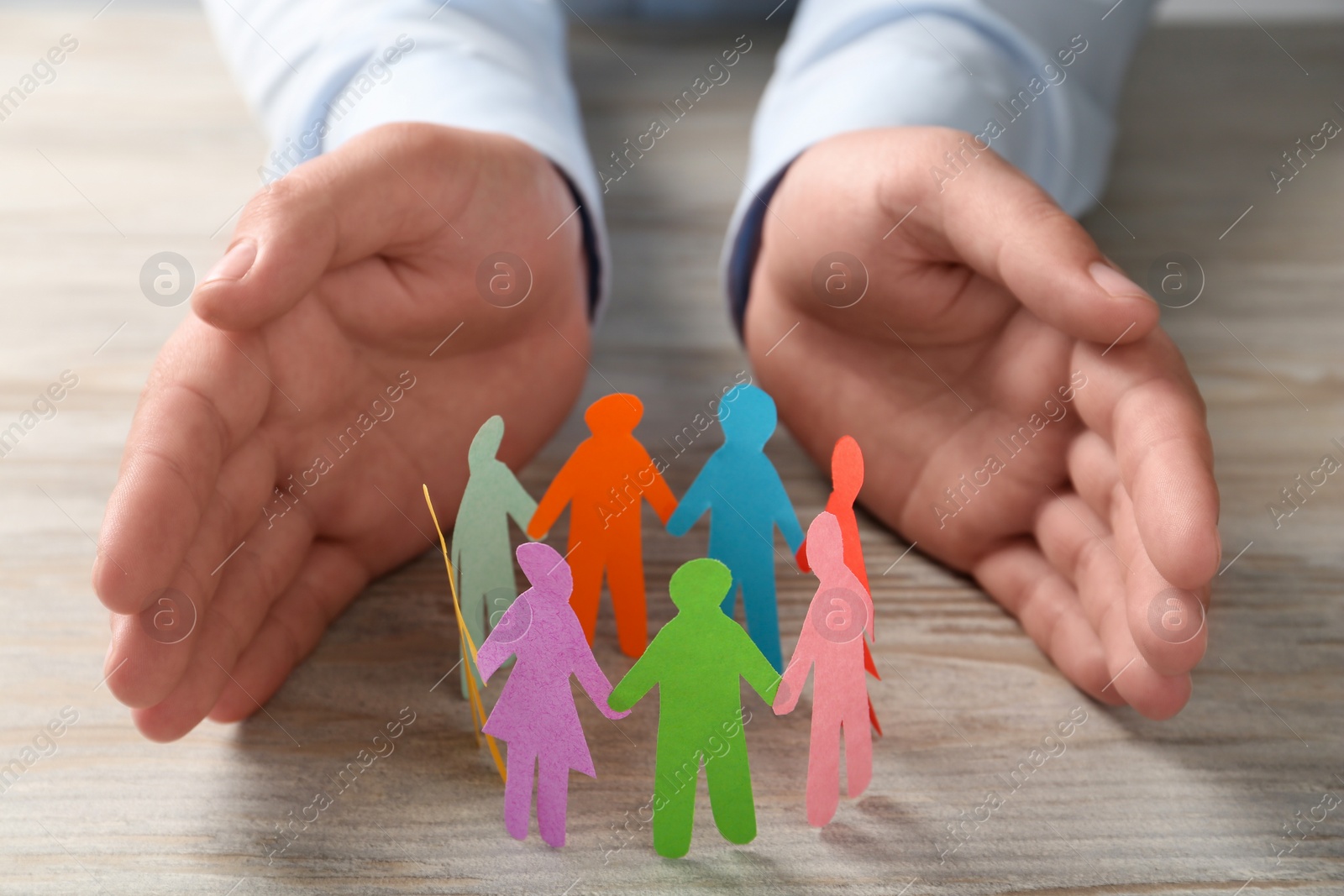 Photo of Man protecting paper human figures on wooden table, closeup. Diversity and inclusion concept